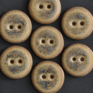 May include: A collection of seven round, two-hole buttons featuring a speckled, mottled glaze in shades of tan, beige, and dark gray. Each button has a smooth, circular edge and a slightly textured surface with a variegated color pattern.