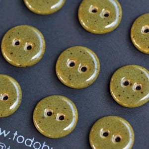 May include: A collection of round, olive green ceramic buttons with a glossy, speckled finish. Each button features two central holes for attachment. The buttons are arranged in a scattered pattern, showcasing their smooth, reflective surface texture.