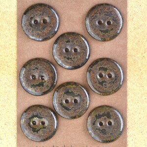 Op de afbeelding: Acht ronde knopen met een gevlekt bruin en groen patroon, elk met twee gaatjes. De knopen zijn gerangschikt op een lichtbruine kaart. De knopen hebben een rustieke uitstraling.