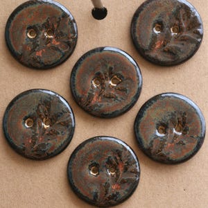 May include: Six handmade ceramic buttons with a glossy finish. The buttons are round, with a dark teal base colour and reddish-brown accents. Each button has two holes for sewing.