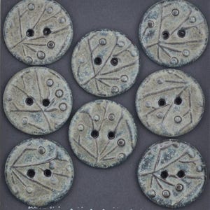 May include: A set of seven circular buttons in a muted grey-green tone with a textured finish. Each button features two central holes and an embossed pattern of radiating lines and small raised dots across the surface of the round discs.