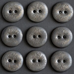 May include: Nine round, silver-toned buttons with two holes each are arranged in a three-by-three grid. The buttons have a textured, floral-like pattern on their surface. The buttons are likely for sewing or crafting.