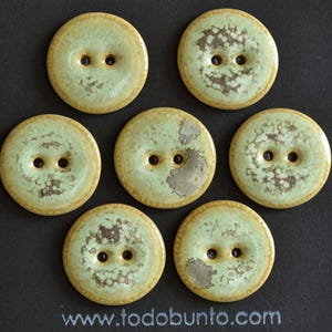 May include: Seven round, ceramic buttons with two holes each. The buttons have a light green center with brown and white speckled patterns and a tan outer edge. The buttons are arranged on a black surface.