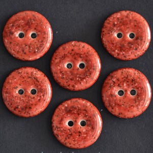 May include: A set of five round, two-hole buttons featuring a glossy, speckled finish in shades of reddish-orange and black. Each button has a smooth, slightly domed surface with two centered holes for attachment, arranged in a cluster.