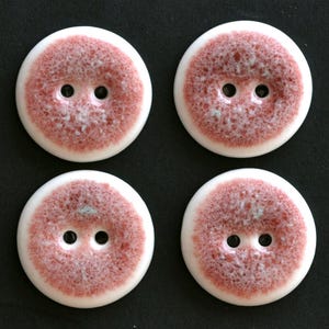 May include: Four round buttons with two holes each, featuring a smooth white outer rim and a textured, mottled reddish-pink center glaze. The buttons have a glossy finish and a circular shape, arranged in a two-by-two grid pattern on a dark background.