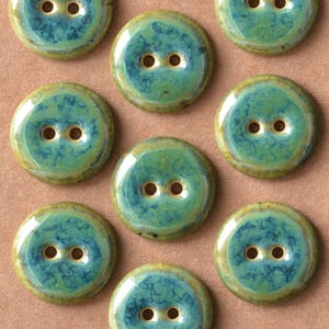 May include: A selection of round ceramic buttons with a glossy finish. The buttons have a teal and green marbled design, two holes, and a gold-coloured rim. They are arranged on a light brown surface.