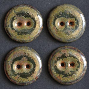 May include: Four circular buttons featuring a glossy, mottled glaze in shades of dark green, olive, and earthy brown. Each button has a smooth, rounded surface with two central holes surrounded by a subtle, warm-toned copper-coloured ring.