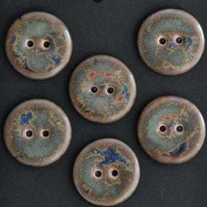May include: Six round ceramic buttons with two holes each. The buttons have a mottled glaze in shades of green, brown, and blue, with hints of orange. The buttons are arranged on a dark surface.