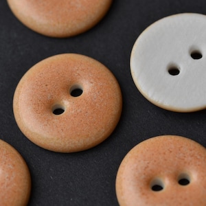 Könnte beinhalten: Nahaufnahme mehrerer runder Knöpfe. Die Knöpfe sind in Beige- und Weißtönen gehalten und haben jeweils zwei oder vier Löcher. Die Knöpfe haben eine glatte, matte Oberfläche. Die Knöpfe sind wahrscheinlich zum Nähen oder Basteln gedacht.