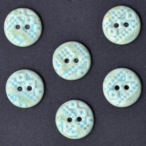 May include: Six round, light blue buttons with a textured, square pattern design. Each button has two holes for sewing. The buttons have a speckled appearance with hints of yellow and are set against a dark background.