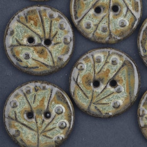 May include: A collection of ceramic buttons with a leaf-like design. The buttons are round with two holes and feature a color palette of green, brown, and gray. The buttons have a textured surface and a glossy finish.