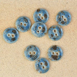 Puede incluir: Un conjunto de nueve botones redondos con un esmalte azul moteado. Cada botón tiene dos o tres agujeros para coser. Los botones tienen un aspecto rústico y hecho a mano, con acentos marrón oscuro.