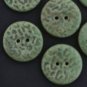 May include: A collection of round, two-hole buttons featuring a textured, mottled surface in shades of sage green and dark brown. Each button displays a unique, uneven pattern across its circular face, with two centered holes for attachment.