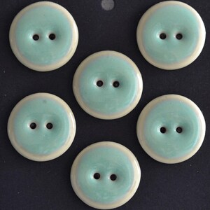 May include: A set of five round, two-hole buttons featuring a glossy mint green centre surrounded by a smooth, cream-coloured border. Each button has a slightly concave surface and a polished finish, arranged in a circular pattern on a dark background.