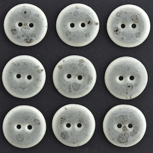 May include: A set of nine round, two-hole buttons arranged in a grid. Each button is white with a translucent, cloudy appearance and features dark grey or black speckled inclusions embedded within the material, creating a unique mottled pattern.