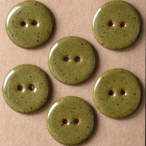 Puede incluir: Un conjunto de cinco botones redondos de color verde oliva con un acabado brillante y moteado. Cada botón presenta dos orificios centrales para su fijación, con una superficie lisa ligeramente abombada y un borde marrón oscuro definido.