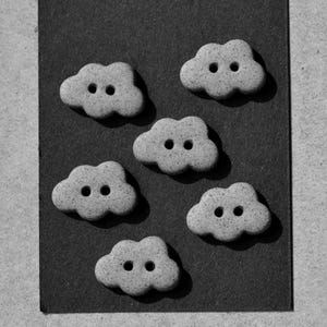 May include: Six cloud-shaped buttons with two holes each, arranged on a dark gray surface. The buttons are a light gray color and have a textured appearance. The image is in black and white.