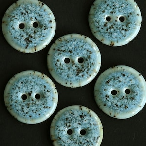 May include: A set of five round, two-hole buttons featuring a glossy, light blue and white speckled glaze with dark brown accents radiating from the centre toward the smooth, cream-coloured outer edges of each individual circular fastener.