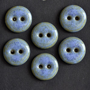 May include: A set of five round, two-hole buttons featuring a mottled glaze finish in shades of periwinkle blue and olive green. Each button has a smooth, glossy surface with a slightly raised center and a recessed area around the two central holes.