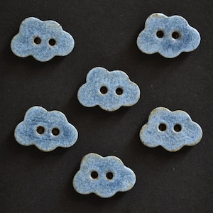 May include: Six cloud-shaped buttons with a speckled blue and white glaze finish. Each button features two central holes for attachment, showcasing a textured surface with variations in color intensity across the individual cloud forms.