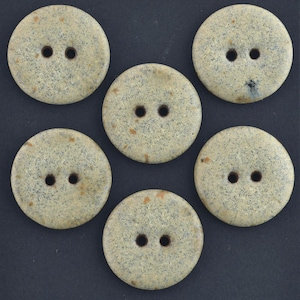 May include: A set of five circular, two-hole buttons with a speckled, light beige and grey textured surface. Each button features a smooth, rounded edge and a consistent pattern of dark grey flecks and subtle tan spots across the front face.