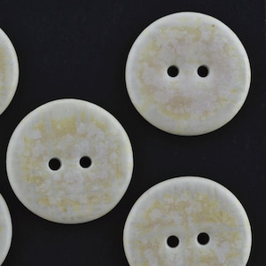 May include: A collection of round, two-hole buttons featuring a mottled, off-white and pale yellow surface texture. Each button has a smooth, circular edge and two centered holes for attachment, displaying a consistent, weathered, and organic appearance.