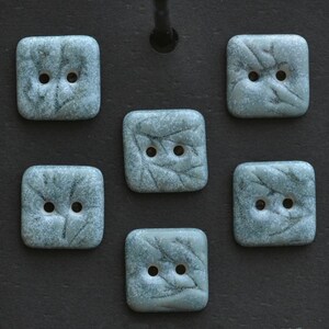 May include: Six square, pale blue buttons with a leaf design and two holes each. The buttons have a speckled texture and are arranged on a dark surface.
