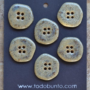 Puede incluir: Seis botones redondos de cuatro agujeros con un acabado esmaltado moteado en tonos tierra marrón y beige. Los botones están dispuestos sobre una tarjeta de presentación gris oscuro con el texto blanco www.todobunto.com y un agujero arriba.