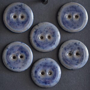 May include: A set of five round, two-hole buttons featuring a glossy, mottled glaze in shades of deep blue, indigo, and soft grey. Each button displays a smooth, circular surface with a distinct speckled pattern and a slightly raised outer edge.