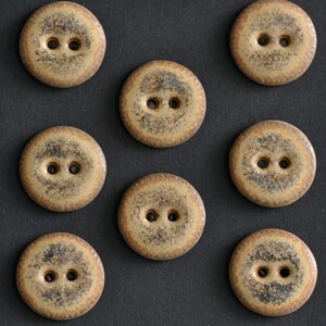 May include: Seven round, two-hole buttons arranged in a grid pattern. Each button features a mottled, earthy tan and dark brown speckled finish with a smooth, slightly glossy surface texture. The buttons are circular with a flat, uniform design.