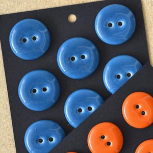 May include: A selection of round, blue buttons with two holes, displayed on a black card. The buttons have a glossy finish and a subtle marbled effect. Some orange buttons are also present.