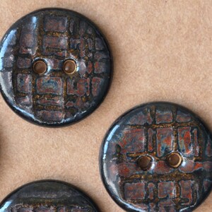 May include: Three round buttons featuring a glossy, dark finish with a textured, grid-like pattern in shades of deep blue, black, and burnt orange. Each button has two central holes for attachment, showcasing a smooth, reflective surface.