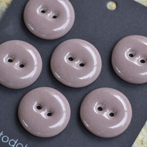 May include: Close-up of six round, mauve-colored buttons with two holes each. The buttons have a glossy finish and are arranged on a dark gray surface. These buttons are ideal for sewing and craft projects.