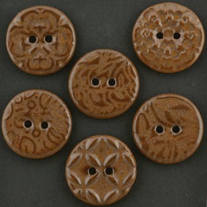 May include: A set of five round, brown ceramic buttons, each featuring a unique embossed pattern on the surface. Every button has two holes in the center for attachment. The buttons have a glossy, glazed finish and are arranged in a circular formation.