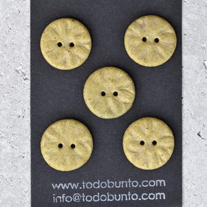 May include: Five round, light green buttons with two holes each, arranged on a dark gray surface. The buttons have a textured, slightly ridged design. The website address and email address are visible at the bottom.