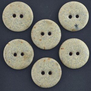 May include: Six round, speckled buttons with two holes each. The buttons are a light beige color with darker speckles and a slightly textured appearance. They are arranged on a dark background.