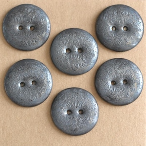 May include: Six round, gray buttons with two holes each. The buttons have a textured surface with a floral pattern. The buttons are arranged on a tan background.