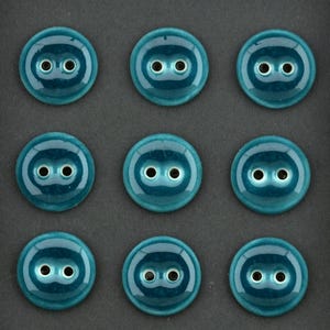 May include: A set of nine round, teal-colored buttons arranged in a three-by-three grid. Each button features a smooth, glossy finish with a recessed center containing two circular holes for sewing, creating a uniform and consistent appearance.