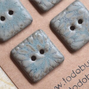 May include: Three square-shaped buttons with two holes each, featuring a textured surface with a mottled grey and teal glaze. The buttons have a raised, petal-like pattern radiating from the center, creating a distinct visual depth and finish.