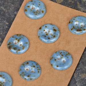 May include: A set of seven round ceramic buttons featuring a glossy light blue glaze with dark brown speckled accents. Each button has two central holes for attachment and a smooth, slightly domed surface, arranged on a brown card backing.