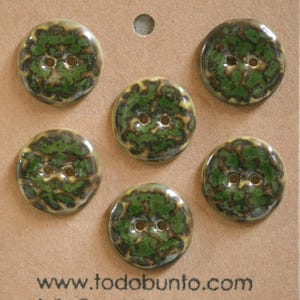 Puede incluir: Cinco botones redondos con acabado brillante, con un patrón moteado en colores verde oscuro, marrón y crema. Cada botón tiene dos agujeros centrales. El texto www.todobunto.com está impreso en negro en la parte inferior de la tarjeta.