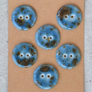 May include: Six round, ceramic buttons with a glossy finish. The buttons are light blue with brown speckles and two holes for sewing. The buttons are arranged on a tan surface.