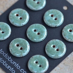 Puede incluir: Un conjunto de ocho botones redondos de dos agujeros con un esmalte brillante de color verde espuma de mar moteado. Los botones están dispuestos sobre una tarjeta negra con el texto blanco todobunto.com, parcialmente visible abajo a la izquierda.