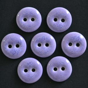 May include: Seven round, two-hole buttons in a light lavender purple color. Each button features a smooth, glossy finish with a subtle, mottled texture that creates a soft, variegated appearance across the surface of the circular fasteners.