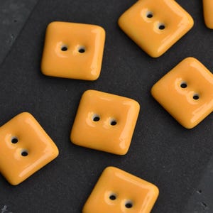 Könnte beinhalten: Nahaufnahme mehrerer quadratischer, orangefarbener Knöpfe mit jeweils zwei Löchern. Die Knöpfe haben eine glänzende Oberfläche und sind auf einer dunklen Oberfläche angeordnet. Diese Knöpfe sind ideal für Näh- und Bastelprojekte.