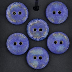 May include: A set of six round, two-hole buttons featuring a mottled blue and gold finish. Each button has a smooth, circular surface with two centered holes for attachment, displaying a variegated color pattern of deep blue and metallic gold.