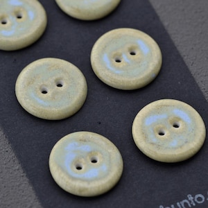 May include: A collection of six round ceramic buttons. Each button has two holes and a light blue and beige color scheme. The buttons are arranged on a dark gray surface.