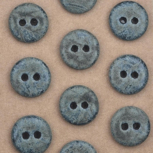 Puede incluir: Colección de botones redondos azul-grisáceos, cada uno con dos agujeros. Los botones tienen un aspecto texturizado y moteado, dispuestos sobre una superficie marrón claro. Adecuados para proyectos de costura y manualidades.