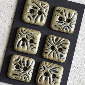May include: Six square, ceramic buttons with a textured, off-white glaze and black, linear designs. Each button has two black holes for sewing. The buttons are arranged on a black surface.