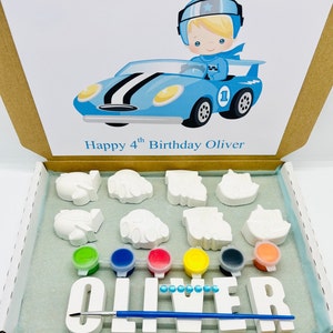 May include: A paint your own craft kit featuring a blue racing car and the words "Happy 4th Birthday Oliver" on the box. The kit contains white ceramic figurines of cars, planes, and a name spelled out in large letters. The kit also includes six small pots of paint and a paintbrush.